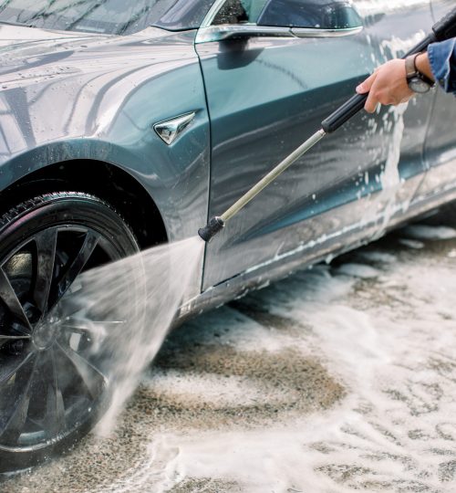 Cropped horizontal shot of young man washing his blue modern electric luxury car, rinsing the soap with high pressure water jet. Manual car wash with pressurized water in car wash outside.