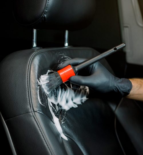 Close up cropped image of washing car interior and seat by a soft brush with foam. Young male worker in black protective gloves cleaning car seat with foam and brush.